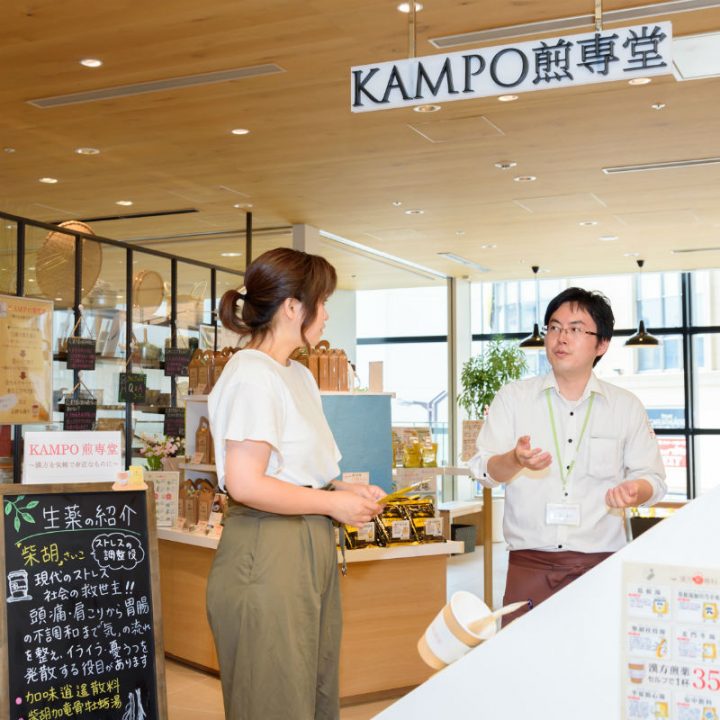 「KAMPO（カンポー）煎専堂」の店頭で男性スタッフから説明を受ける女性