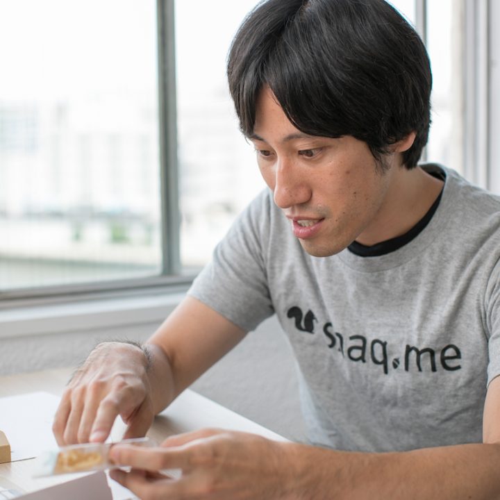 お菓子を指さして説明するグレーのTシャツを着た「snaq.me」代表取締役の服部慎太郎さん