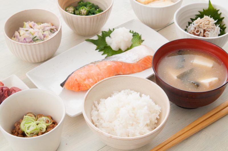 和朝食の写真。ご飯、豆腐の味噌汁、焼き鮭、納豆などがならんでいる