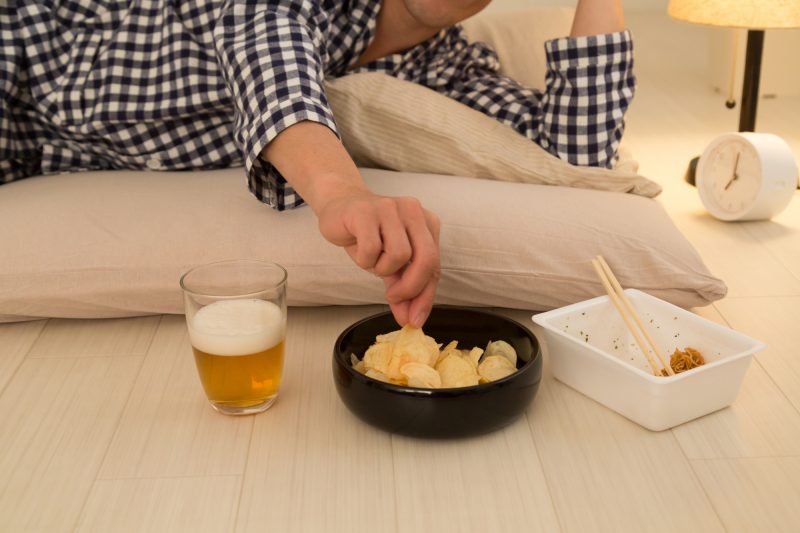 男性が布団に寝ながらビールを飲み、ポテトチップスに手を伸ばし、食べ終わったカップ焼きそばが置いてある