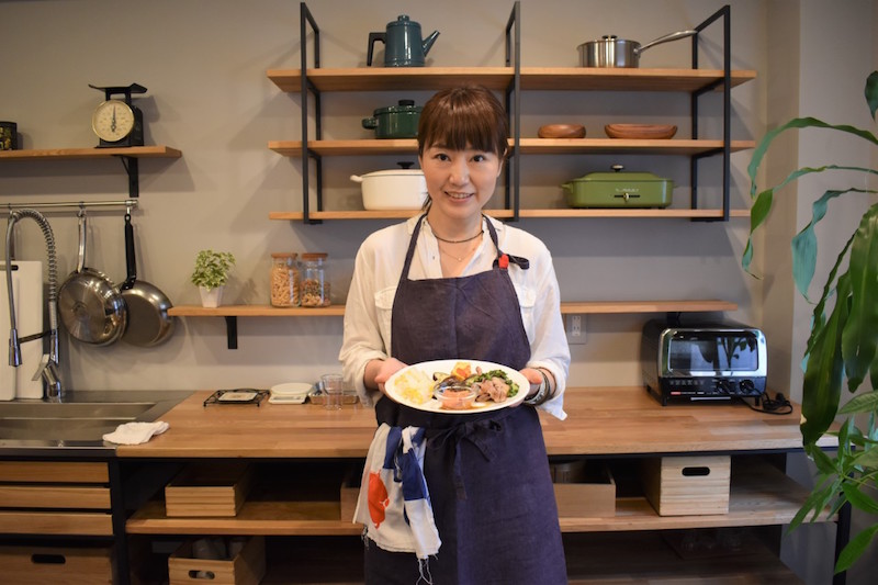 料理家の井上かなえさんが料理を盛った皿を両手で持ってキッチンに立っている