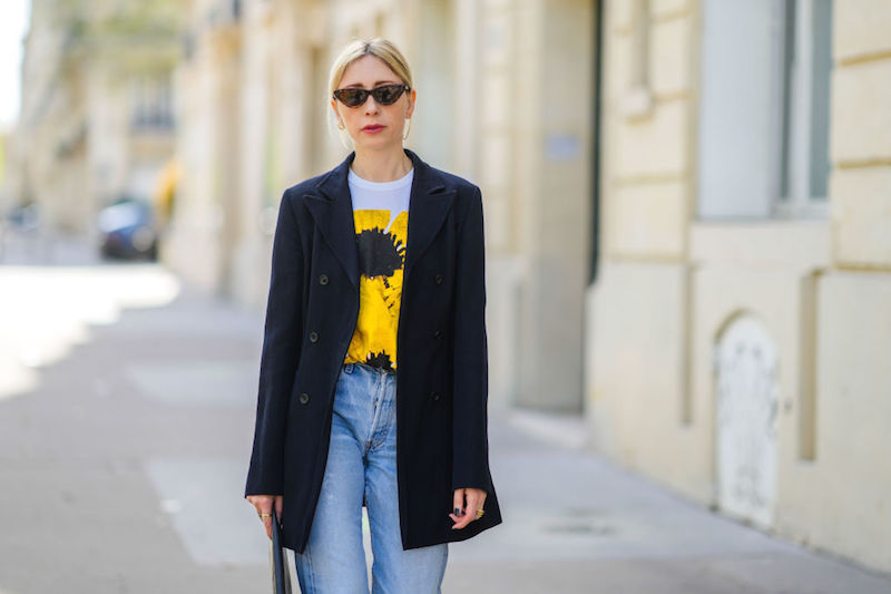 白のひまわり柄のTシャツにデニムパンツ、黒のジャケットを羽織サングラスをかけた女性