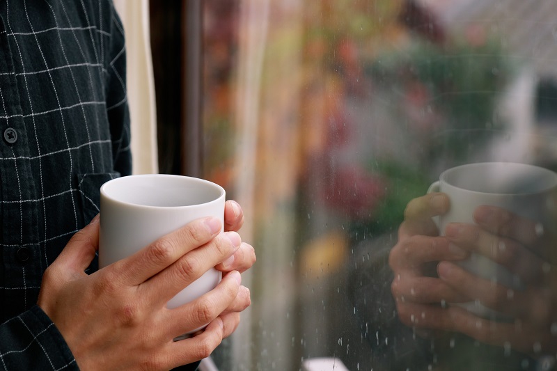 窓際でマグカップを手に雨降る外を眺める人の手元画像