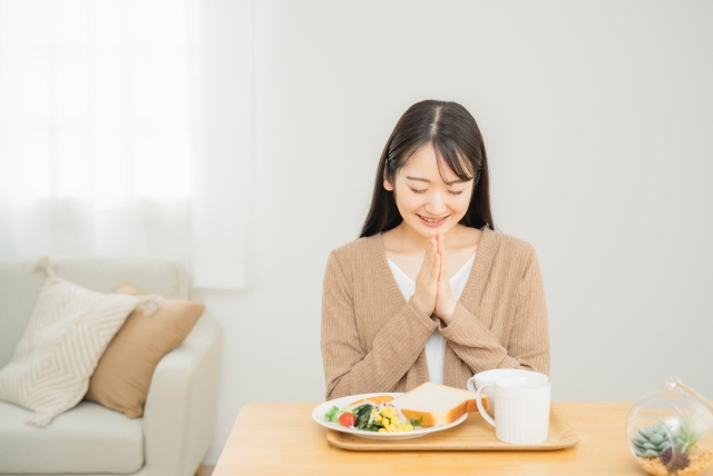 食パンやサラダが乗ったお皿やマグカップをのせたテーブルを前に、手を合わせいただきますをする女性