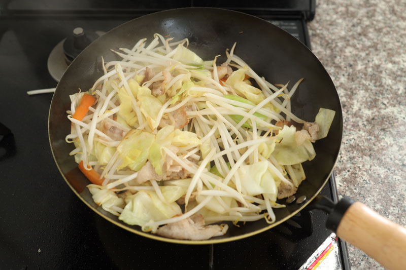 フライパンでごま油を中火で熱する。豚バラ肉、にんじん、キャベツ、もやしの順に加えて炒める