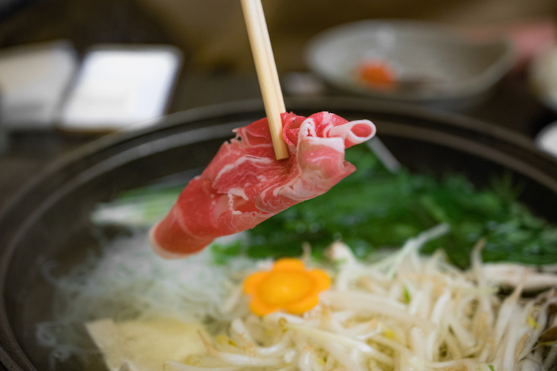 もやしなど野菜が入った鍋に、箸でつまんだ肉を入れようとしている手元