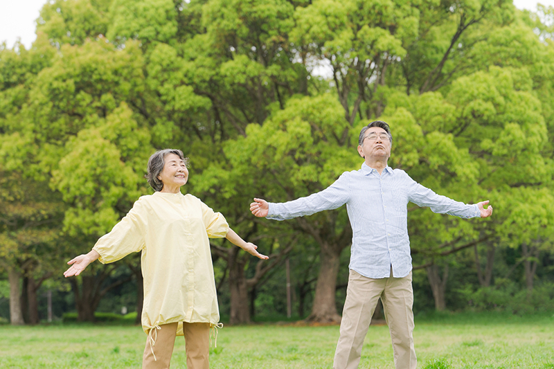 緑のあるところで、両手を広げ深呼吸しているシニア夫婦