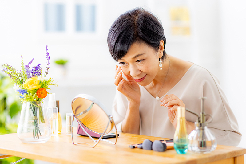 香水やメイクブラシが並ぶテーブルの上の鏡を覗き込み肌をチェックする女性