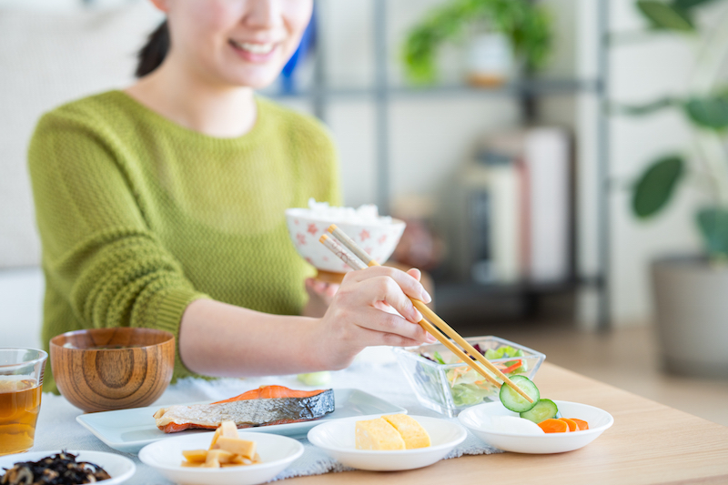 ご飯茶碗を左手に持ち、右手の箸で漬物を取ろうとしている女性の鼻から下
