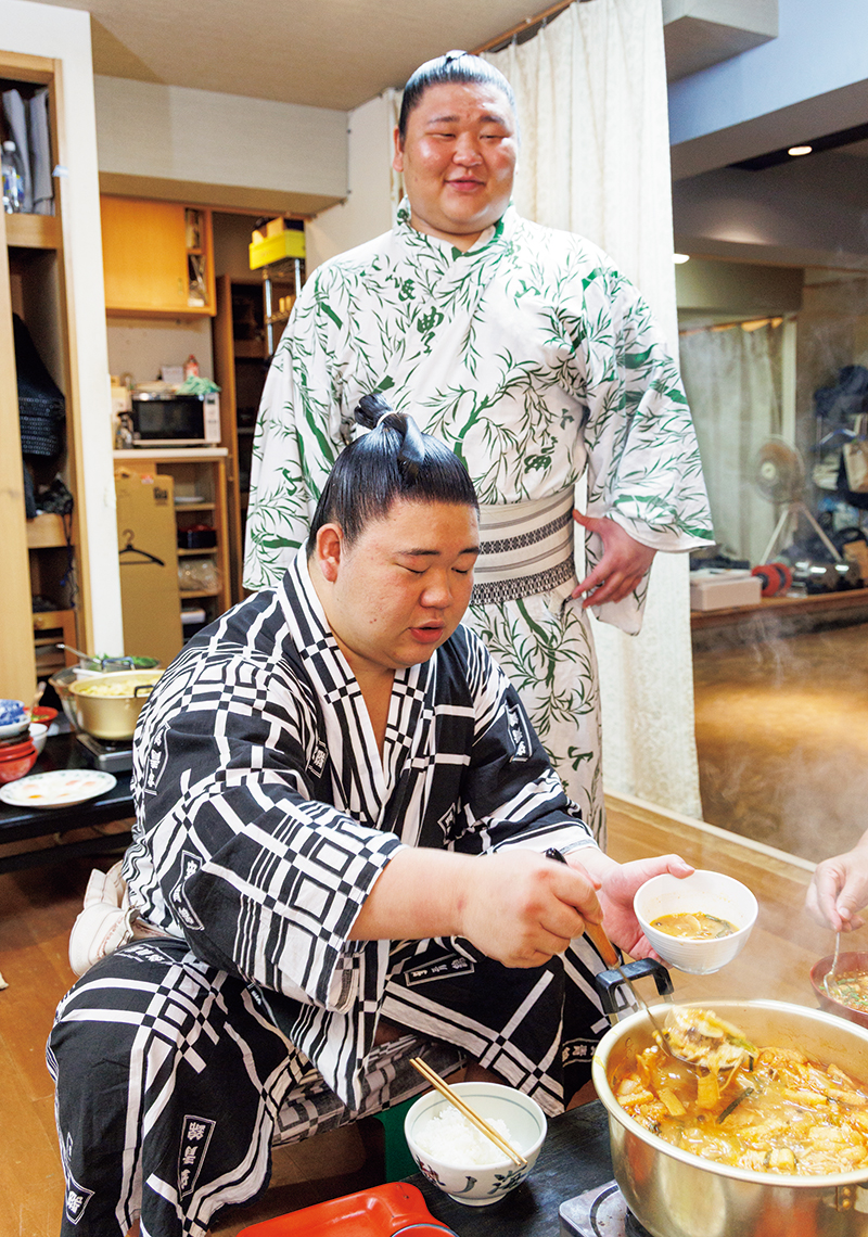 「今日もおいしいです。ご飯がすすみます！」とモリモリ食べる藤ノ川。後ろに立つ弟の幕下・碇潟（いかりかた）は五月場所で幕下優勝を果たし、兄弟ともにいま注目の力士だ。