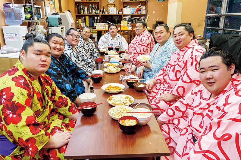 関取はじめ、みな一緒にいただきます！ちゃんこ場の長机を囲んで食事。白飯のほか、雑穀ご飯も用意されているところが、体にやさしい健康ちゃんこを目指す中村部屋らしい。